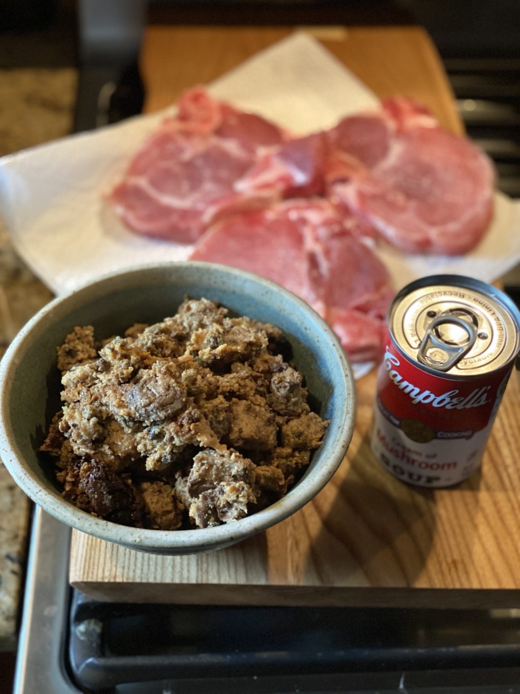 A bowl of stuffing next to a can of Campbell's Cream of Mushroom soup with raw pork chops in the background.