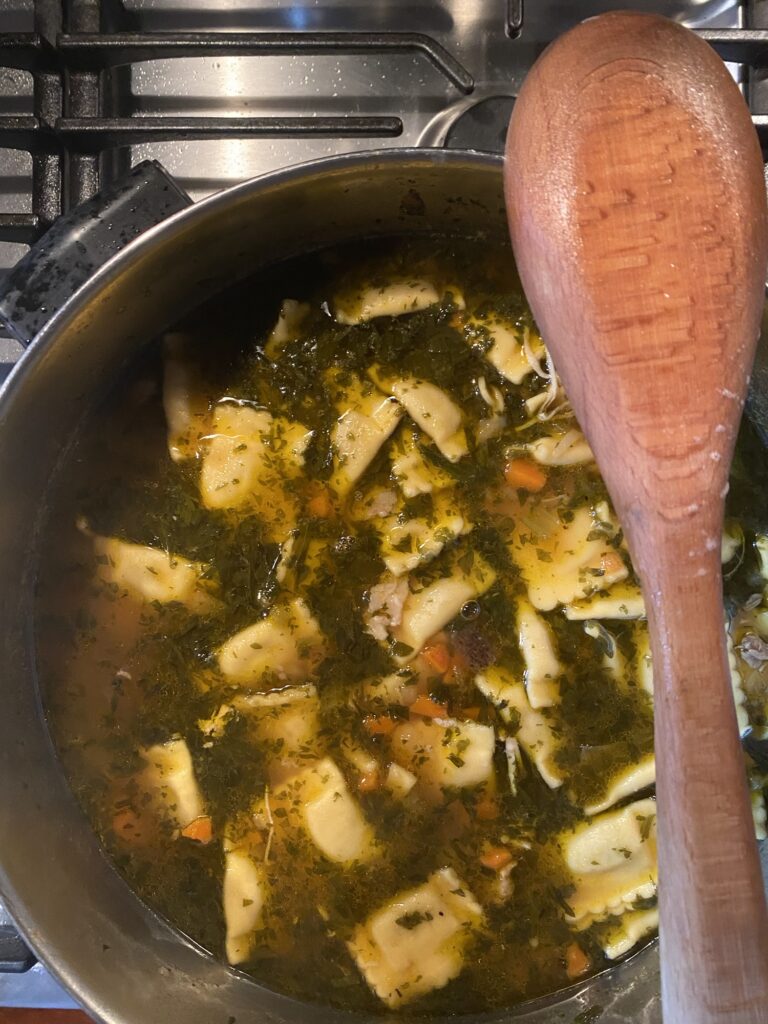 A wooden ladle being held over a pot of chicken soup with ravioli pasta in it.