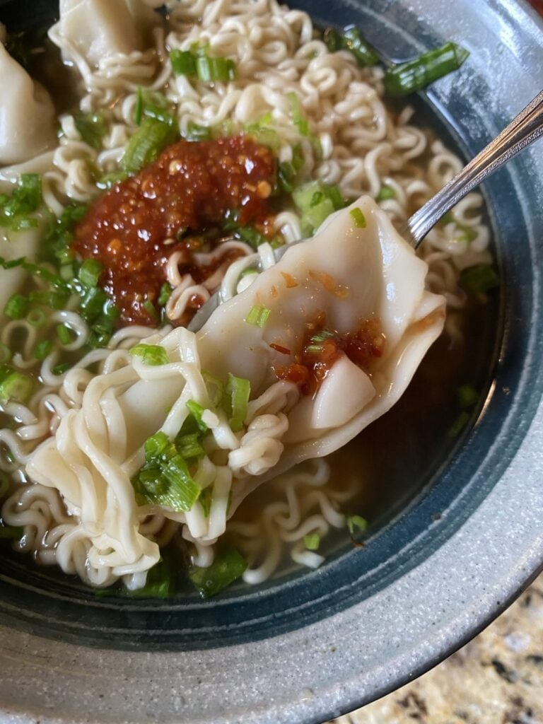 A bowl of noodle soup with dumplings, one dumpling held over the bowl on a spoon.