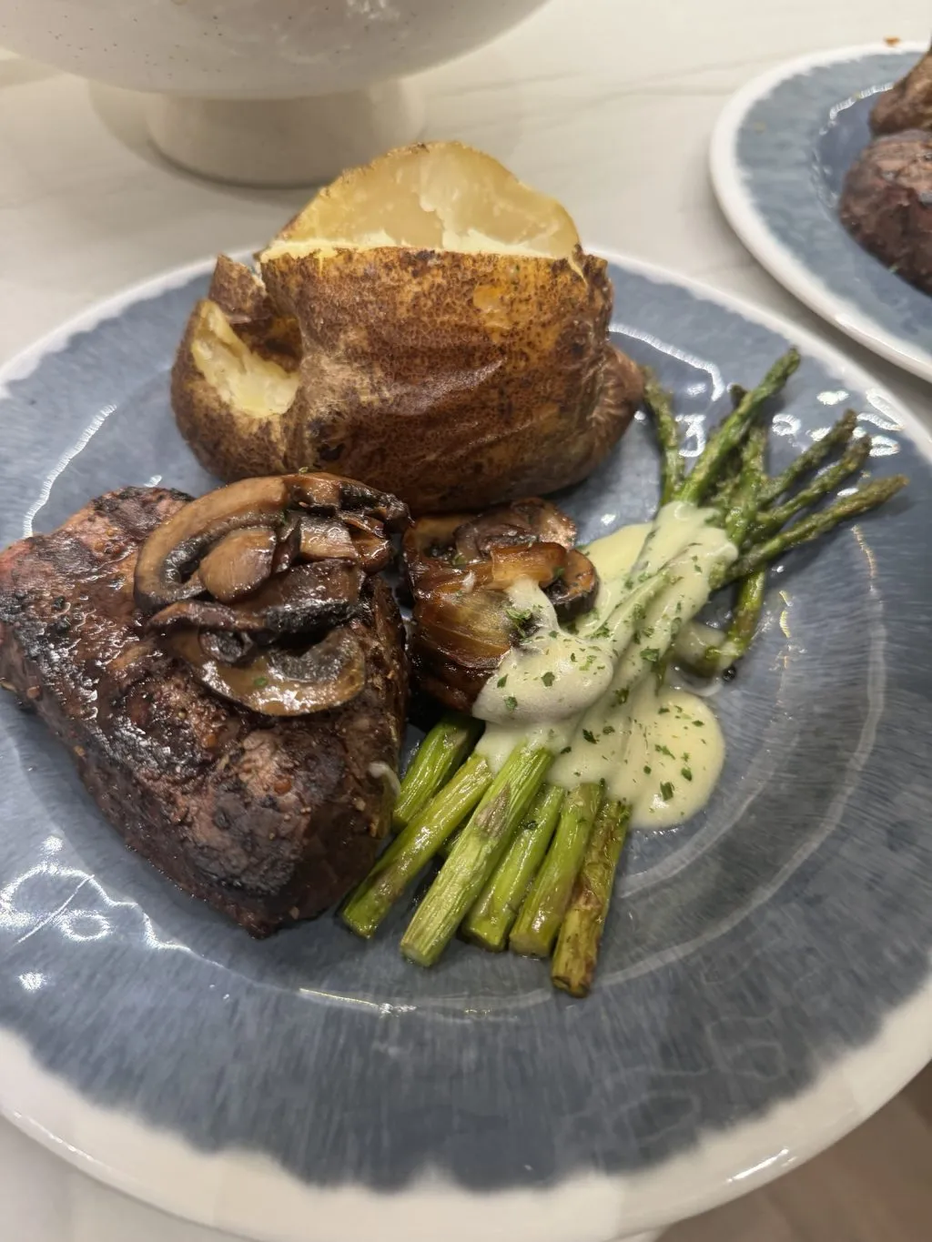 A beef filet, mushrooms, baked potato, and asparagus topped with mushroom sauce on a blue plate