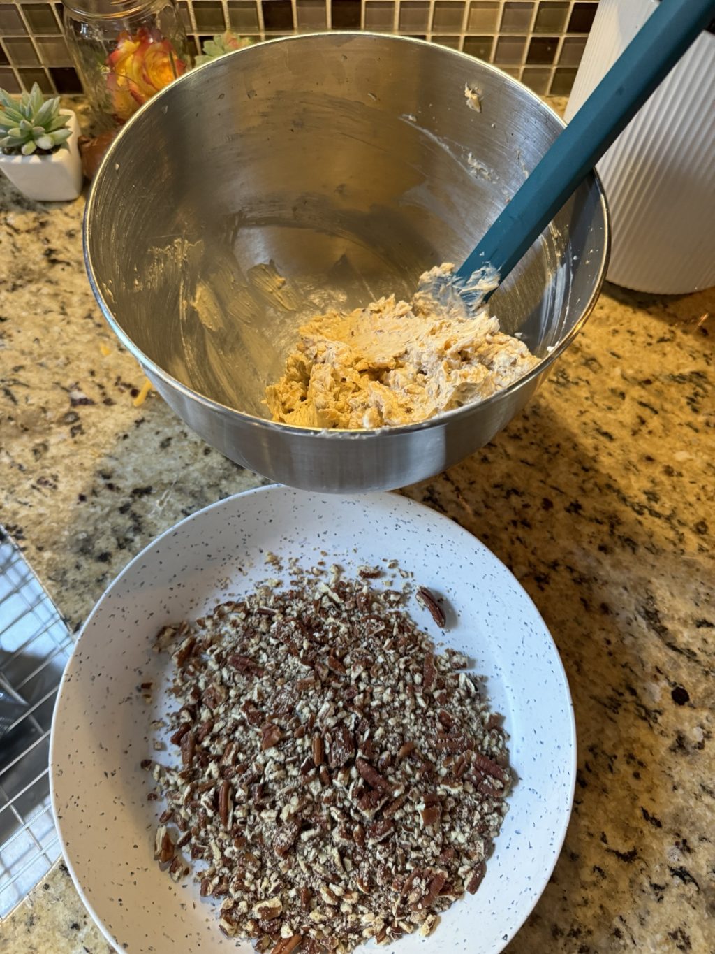 The cheese ball mixture in a metal bowl with a blue spatula held over a white bowl of crushed pecans.