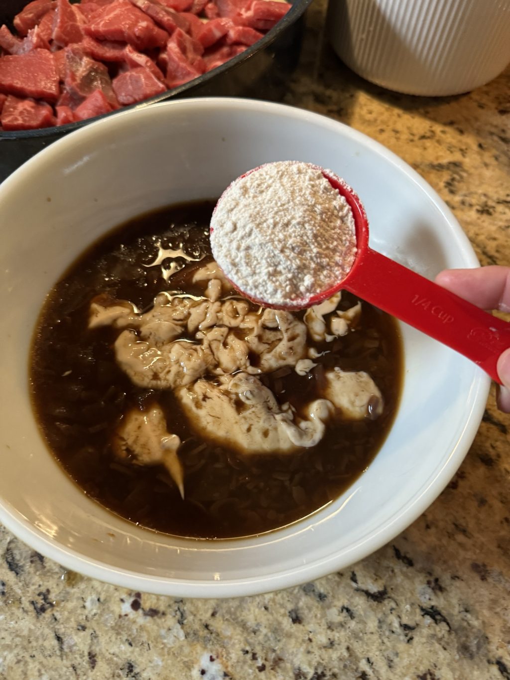 Flour in a red measuring spoon held over a gravy mixture in a bowl.