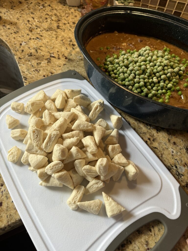 Cut up biscuit dough on a white cutting board next to a specked roaster filled with meat, gravy, and green peas.