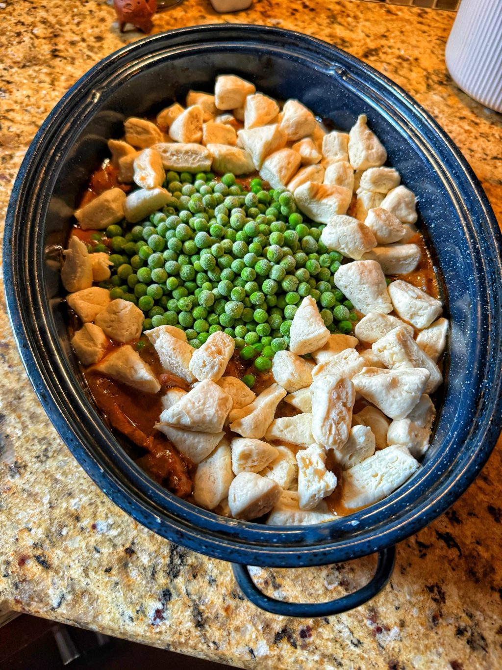 Frozen peas surrounded by raw biscuit dough pieces over beef and gravy in the speckled roaster.