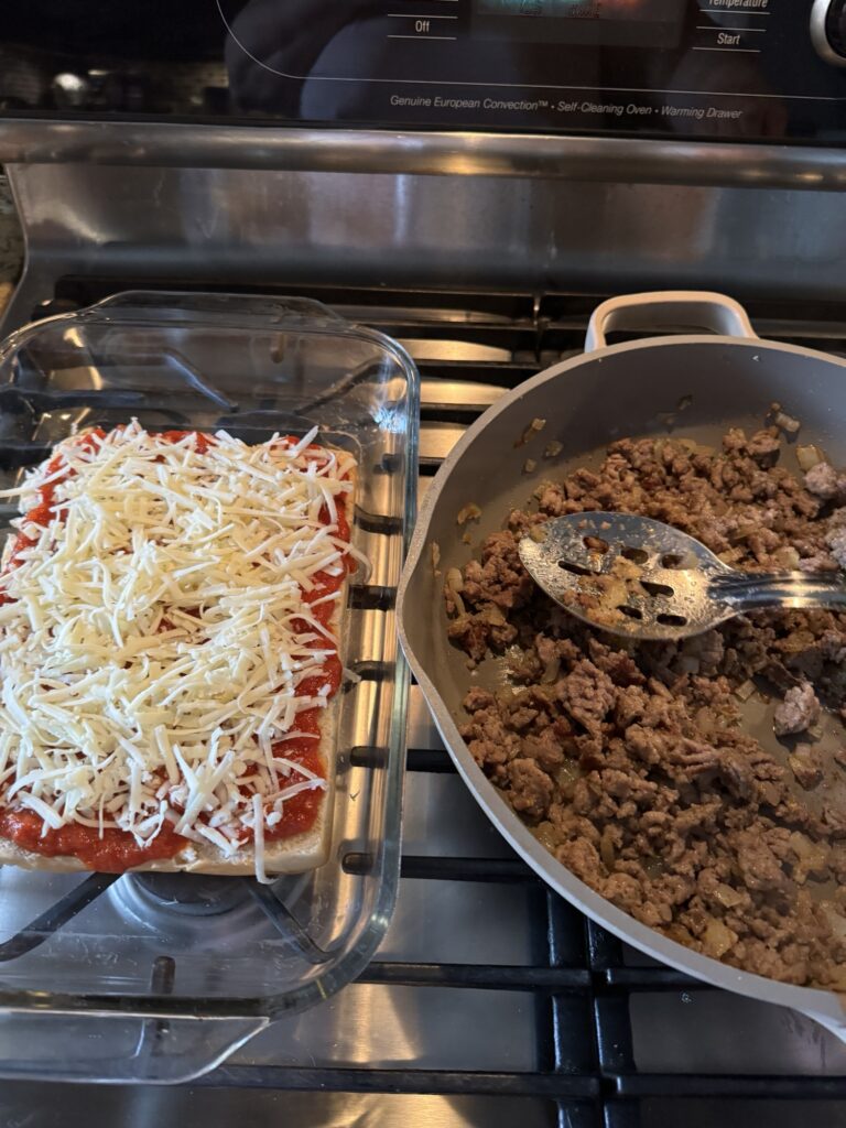 A half slab of rolls topped with marinara sauce and shredded cheese next to a pan of crumbled sausage