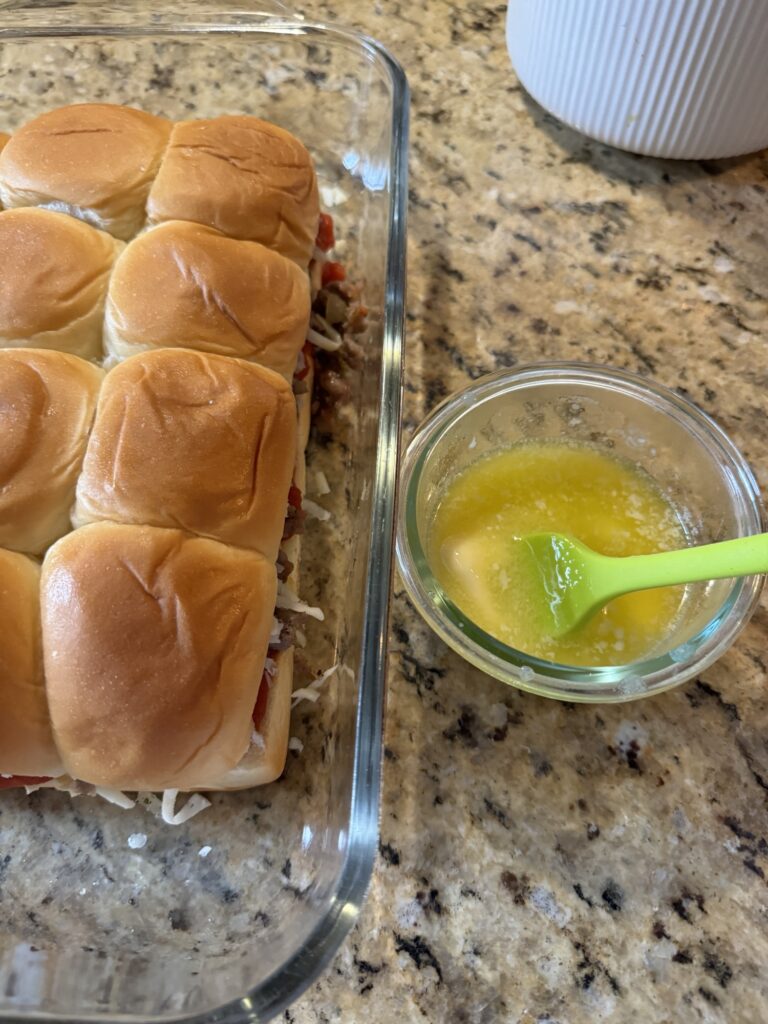 A glass baking dish of the prebaked sliders next to a glass cup of melted butter