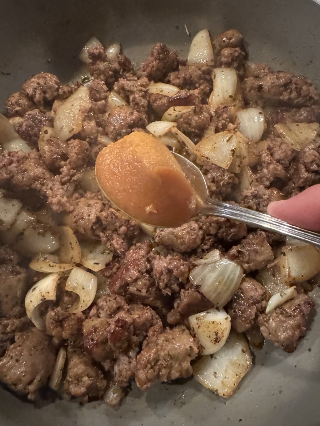 A spoon of bouillon about to be dropped into sausage and onions cooking in a skillet