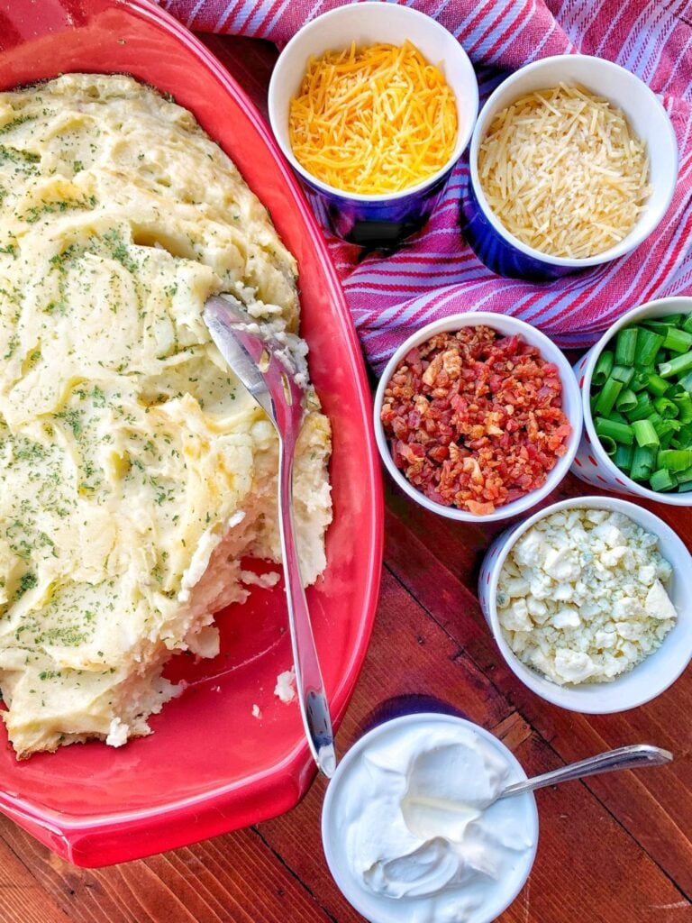 Mashed potato casserole with "fixins" on the side, including cheeses, green onion, sour cream, and bacon bits