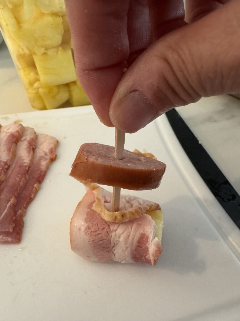 A skewer being put into uncooked bacon wrapped pineapple and a slice of kielbasa