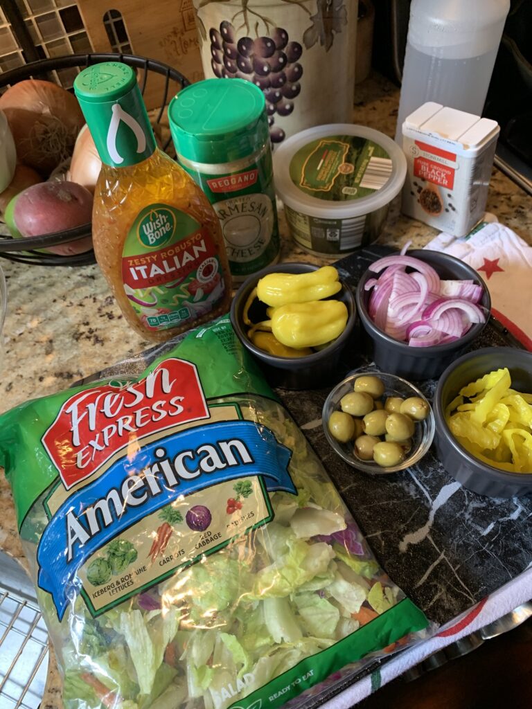 The ingredients for the salad, including a bag of American salad mix, olives, banana peppers, red onion, Italian dressing, and peperonncini