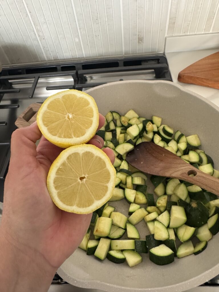 A lemon sliced in half held over zucchini in the pan