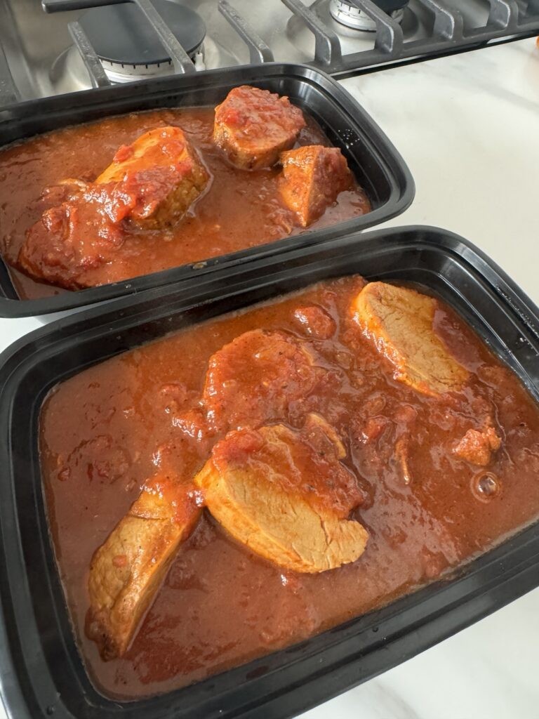 Pork medallions in red sauce in black plastic takeaway containers