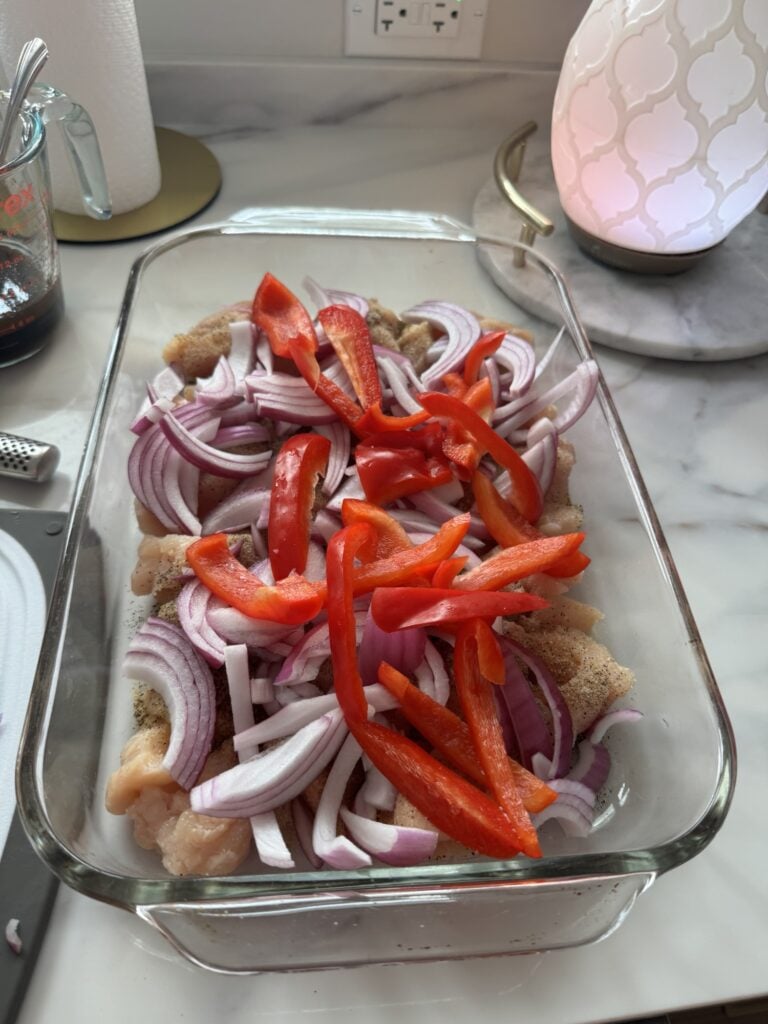 Red peppers and onions over raw chicken cubes in a glass baking dish