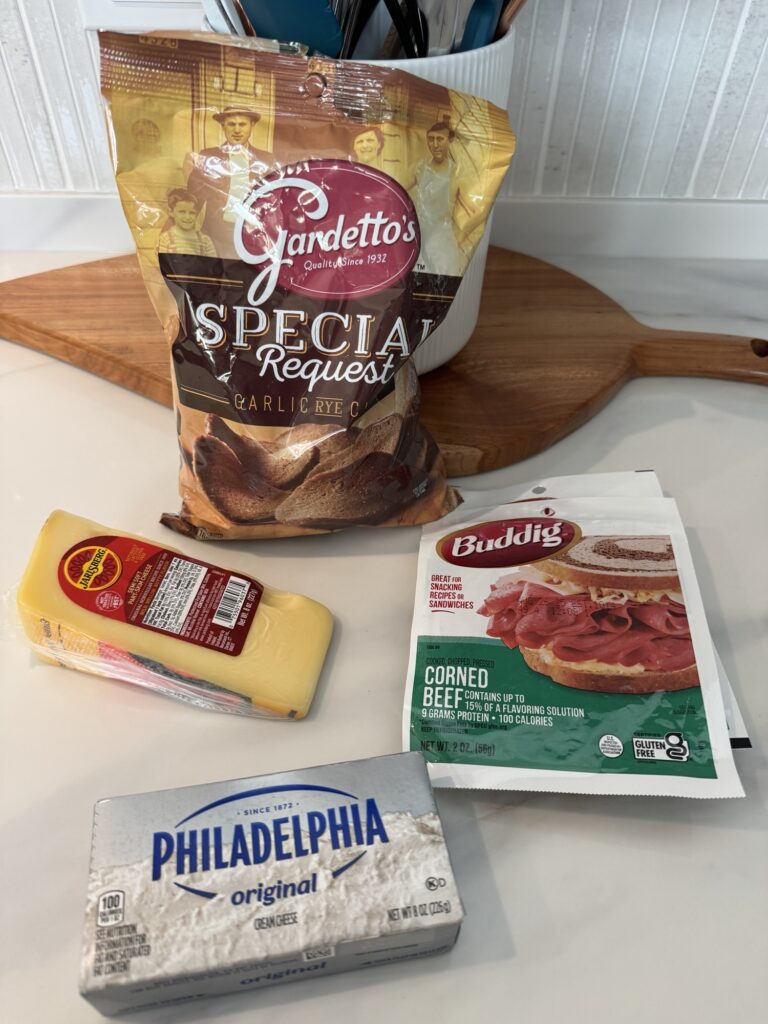 A bag of Gardetto rye chips, a wedge of Jarlsberg cheese, and a package of Buddig corned beef