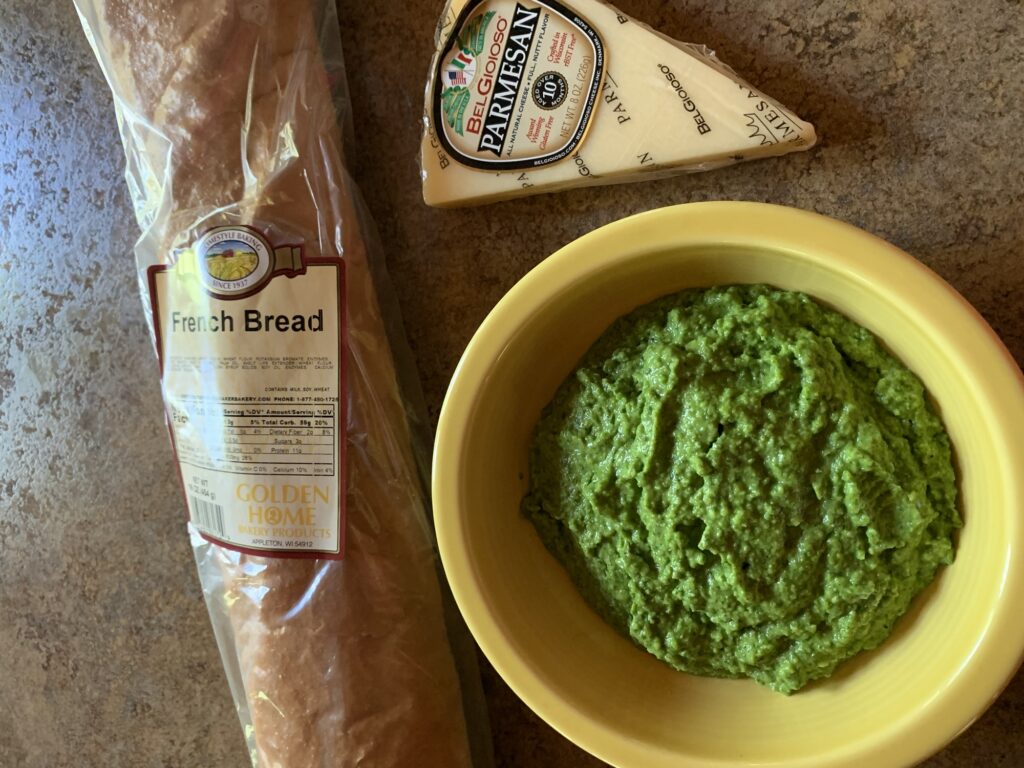 A loaf of French bread, bowl of mushed pea mix, and a wedge of Parmesan cheese