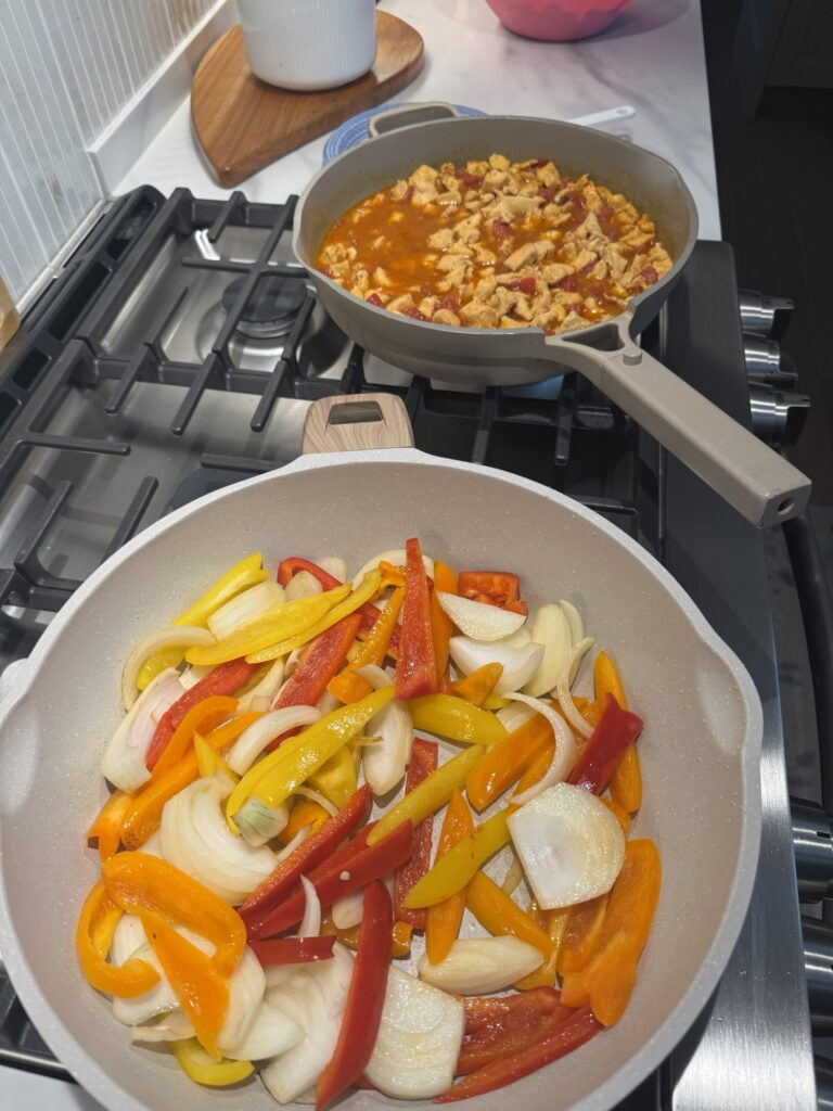 Peppers and onions on the stove next to chicken and sauce on the stove