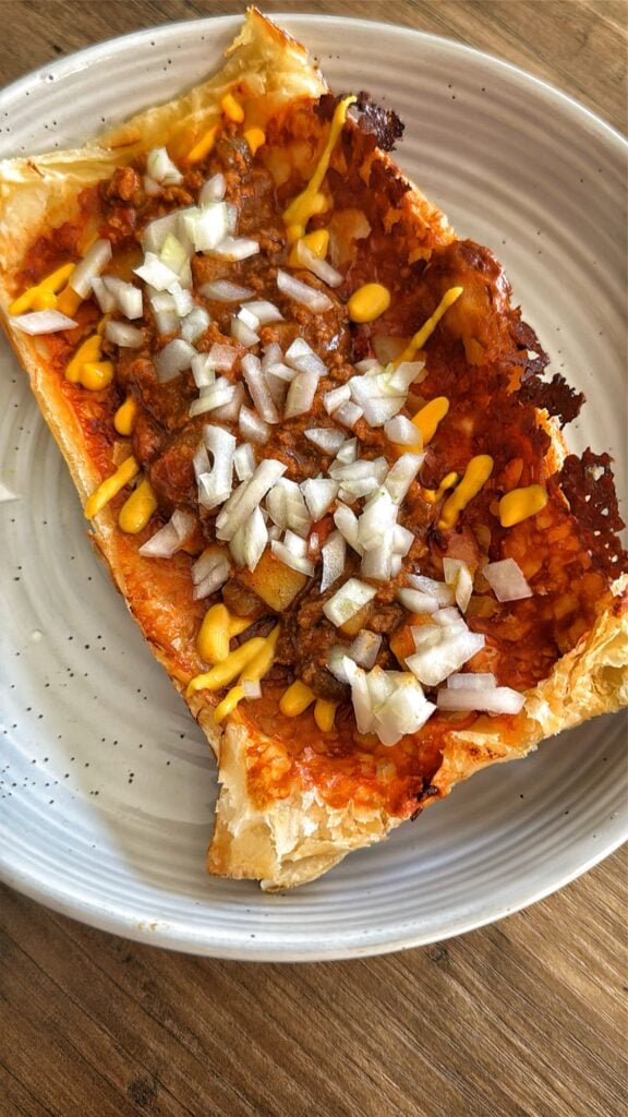 The baked pastry dog boat tipped with chili, mustard, and diced onion